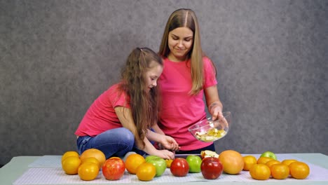 mother with daughter with fruit in the kitchen. young woman and a little child in pink t-shirts prepare fruit salad at home. healthy food.
