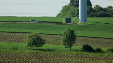 Coche-Estacionado-Junto-A-Una-Turbina-Eólica-En-Un-Soleado-Paisaje-Rural,-Campos-Verdes,-Día-Sereno,-Líneas-Eléctricas-Distantes