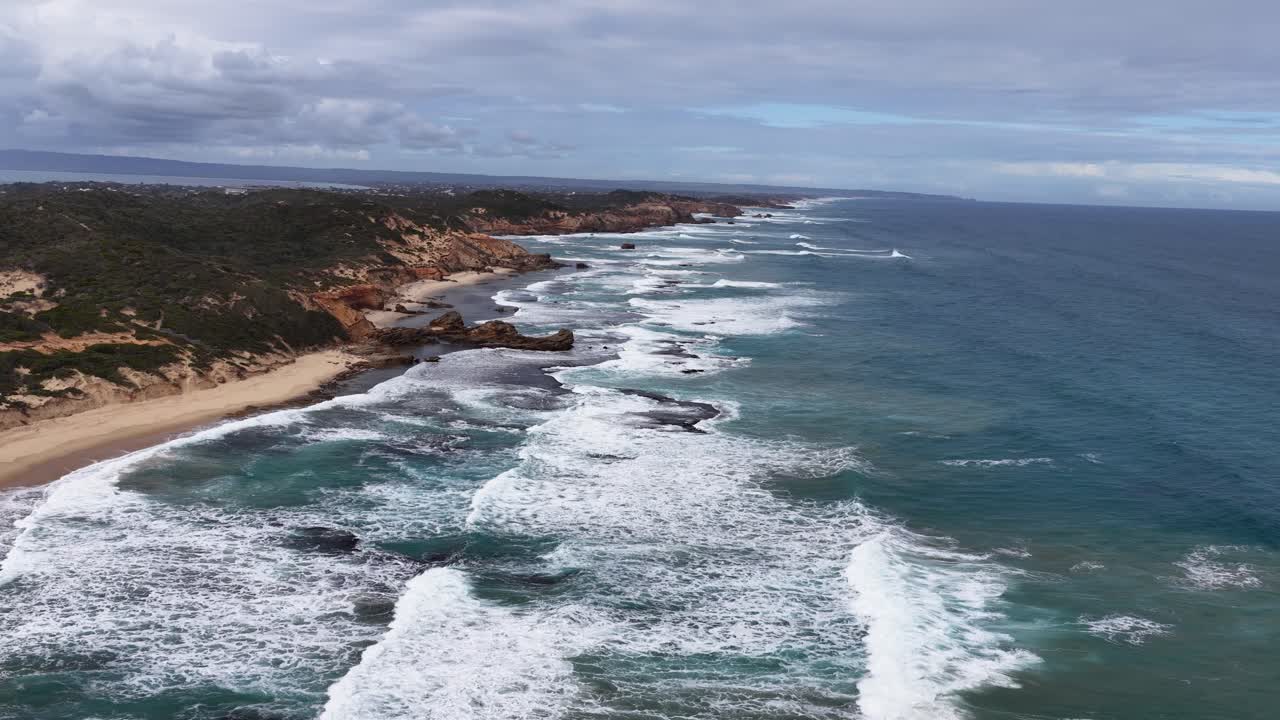 Drone glides above rocky shoreline, capturing turquoise waves, sandy beach, and dramatic coastal cliffs