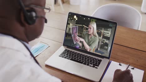 African-american-male-doctor-wearing-phone-headset-taking-notes-while-having-a-video-call-on-laptop