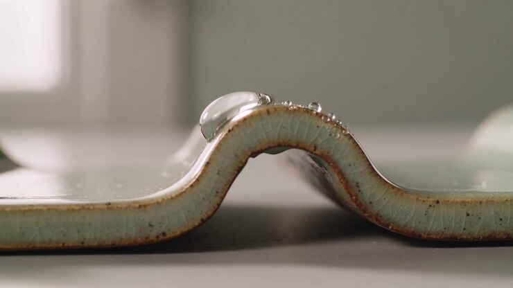 Sliding droplet rightward on wavy ceramic strip on table, shedding beads under gravity, capillarity