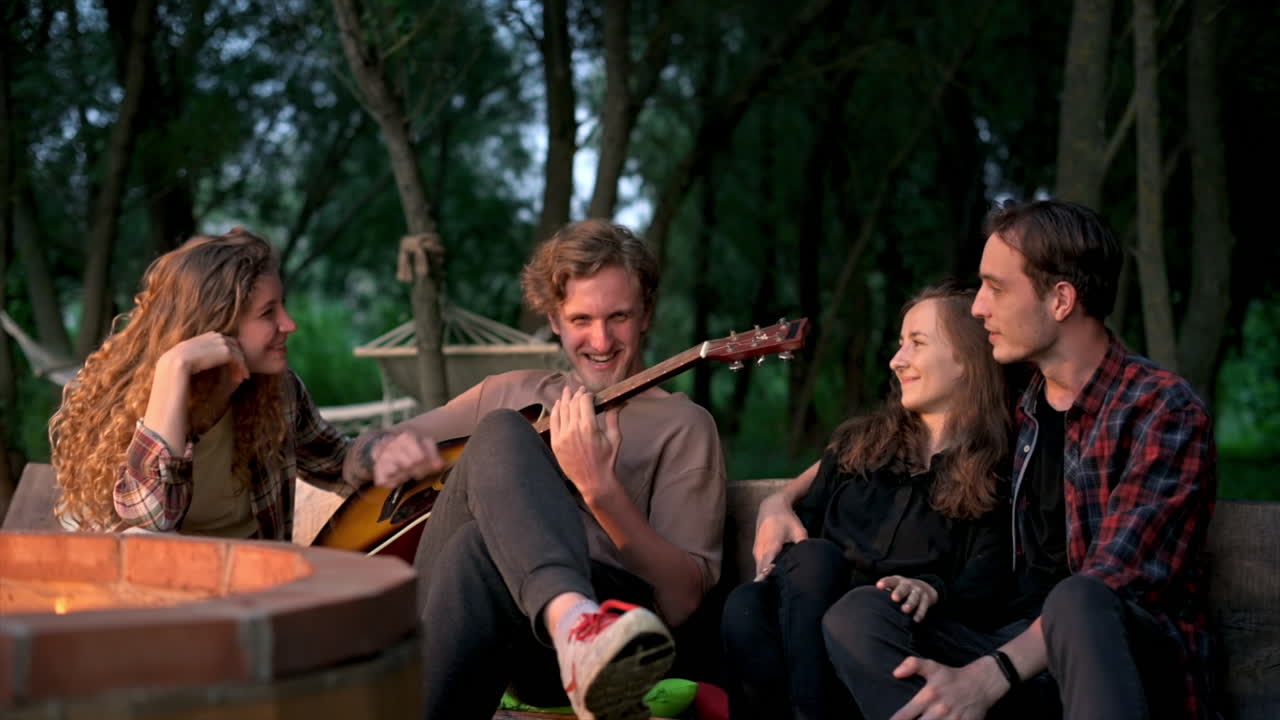 En gruppe glade, unge venner ved et lejrbål under glamping om natten. To mænd og kvinder. Der spiller guitar. Slow motion.