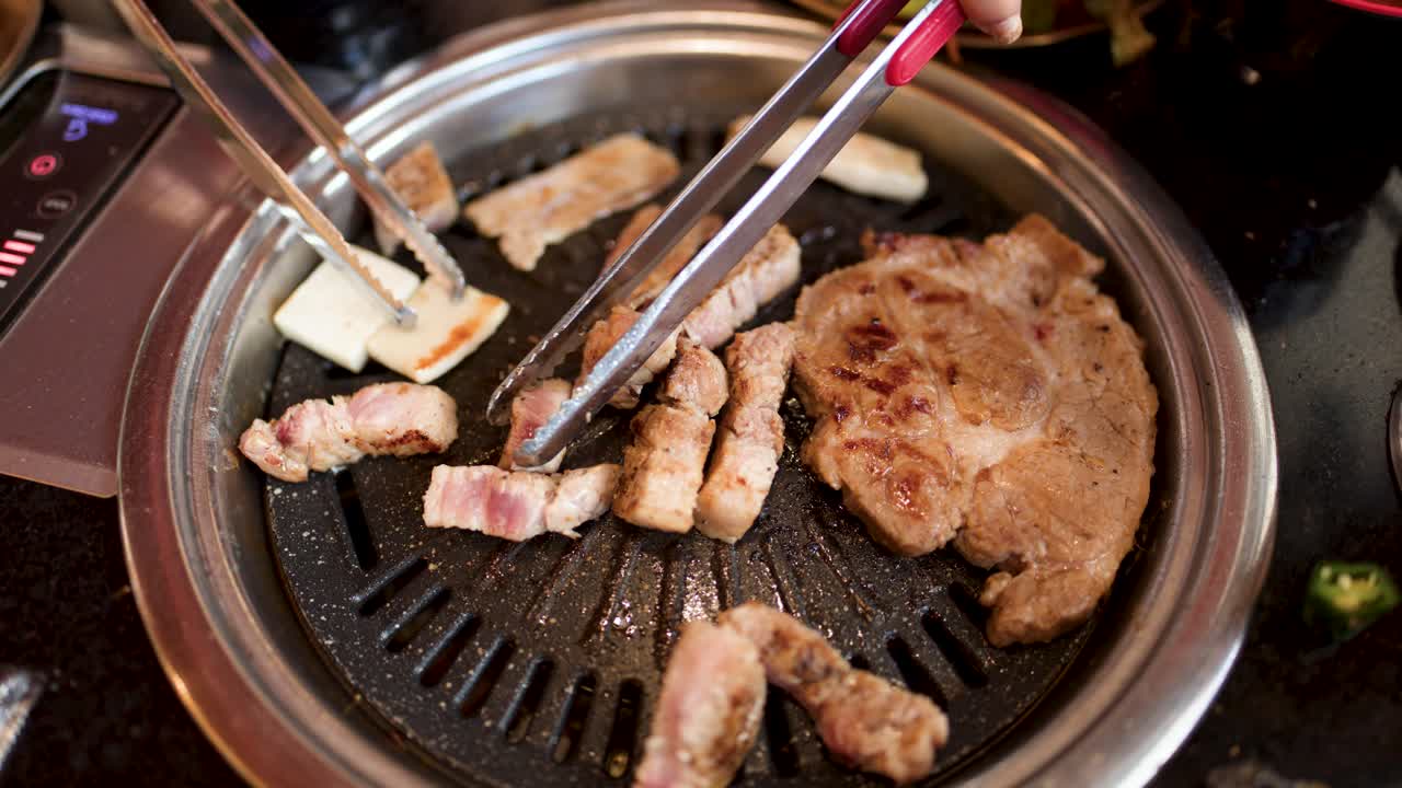 Hand uses tongs to flip pork belly slices on tabletop Korean barbecue grill, warm lighting