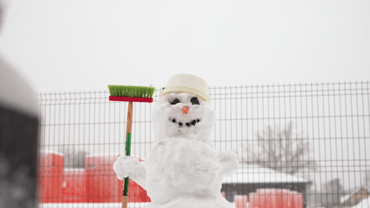 A delightful snowman with a carrot nose and coal eyes stands cheerfully in a snowy backyard, surrounded by bare branches and fenced perimeter. Overcast sky enhances winter atmosphere
