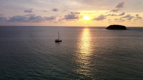 vuelo aéreo sobre las tranquilas aguas del atardecer con vista inclinada hacia arriba de la silueta del yate flotando con la isla en segundo plano