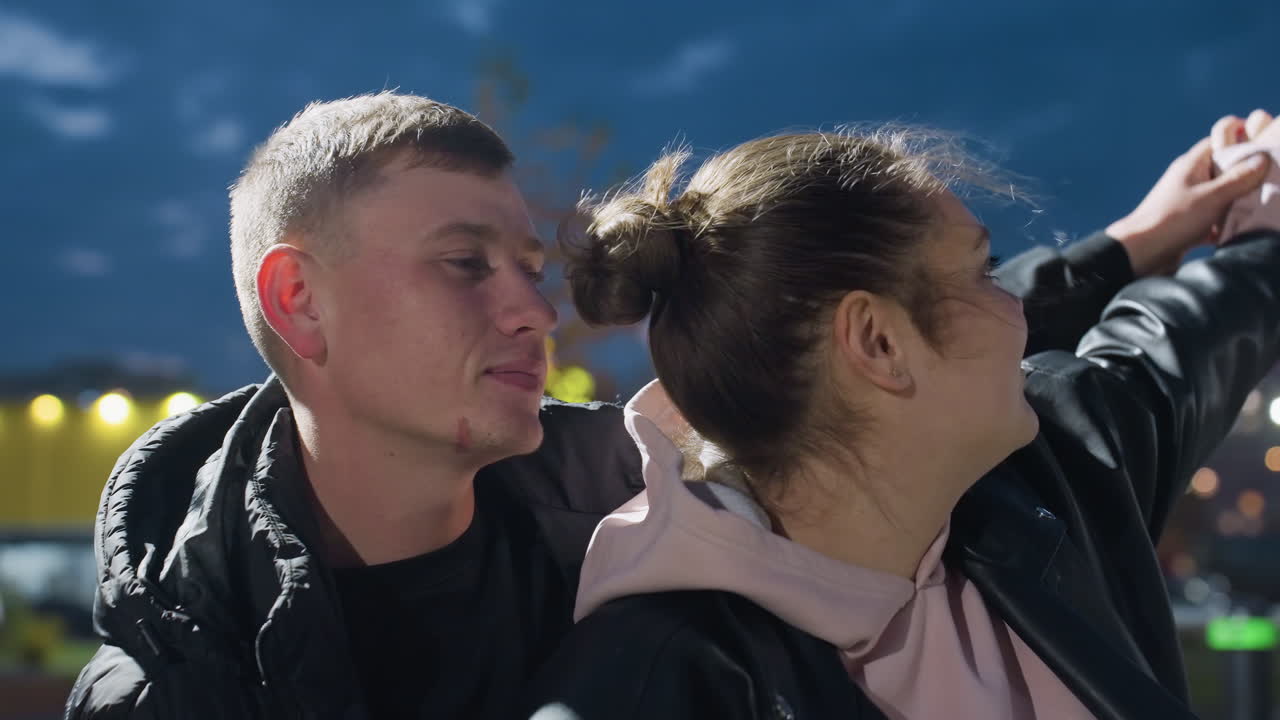 Lovely friends dancing outdoors during windy evening with hair blowing gently, surrounded by glowing residential lights and vibrant urban background reflecting joy