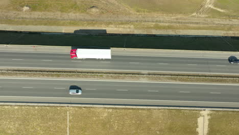 Luftaufnahme-Von-Oben-–-über-Einer-Autobahn,-Der-Starke-Kontrast-Zwischen-Der-Kargen-Landschaft-Und-Den-Fahrzeugen:-Ein-Einzelnes-Auto-Und-Ein-Roter-Lastwagen,-Die-Die-Stille-Und-Bewegung-Des-Transports-Hervorheben