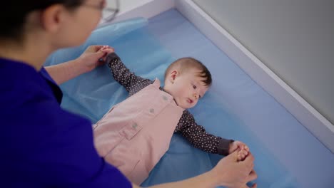 From-above-over-the-shoulder-a-confident-brunette-girl-doctor-in-glasses-finds-a-common-language-and-checks-a-little-baby-girl-in-a-pink-overalls-for-the-correct-development-in-a-modern-clinic-at-a-pediatricians-appointment