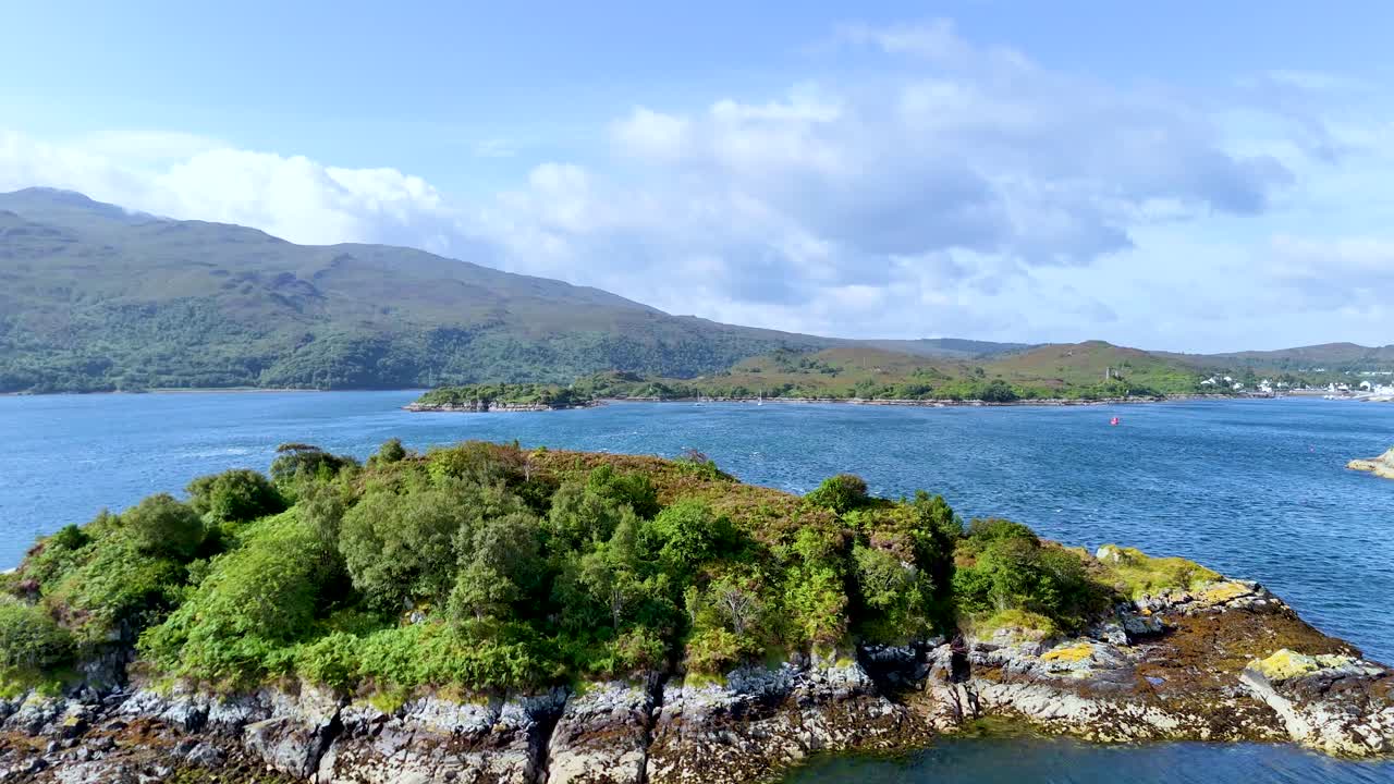 Drone camera smoothly flies over a small, lush island in Loch Alsh, revealing rugged shoreline, blue water, and distant Scottish Highlands under bright daylight