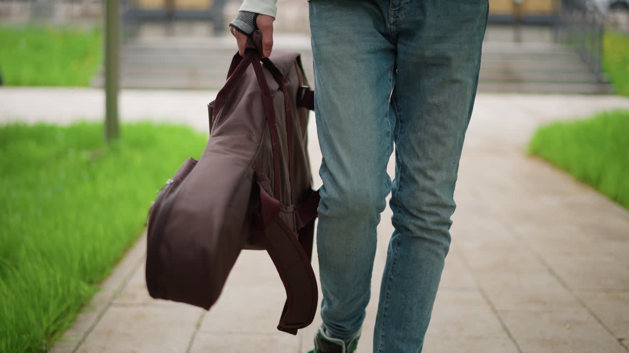 Un chitarrista cammina con la custodia lungo il sentiero del parco, indossa jeans e un maglione casual, la borsa imbottita per chitarra è appesa al suo fianco, prato verde e gradini di pietra sullo sfondo, passo rilassato e sicuro, stile documentaristico spontaneo
