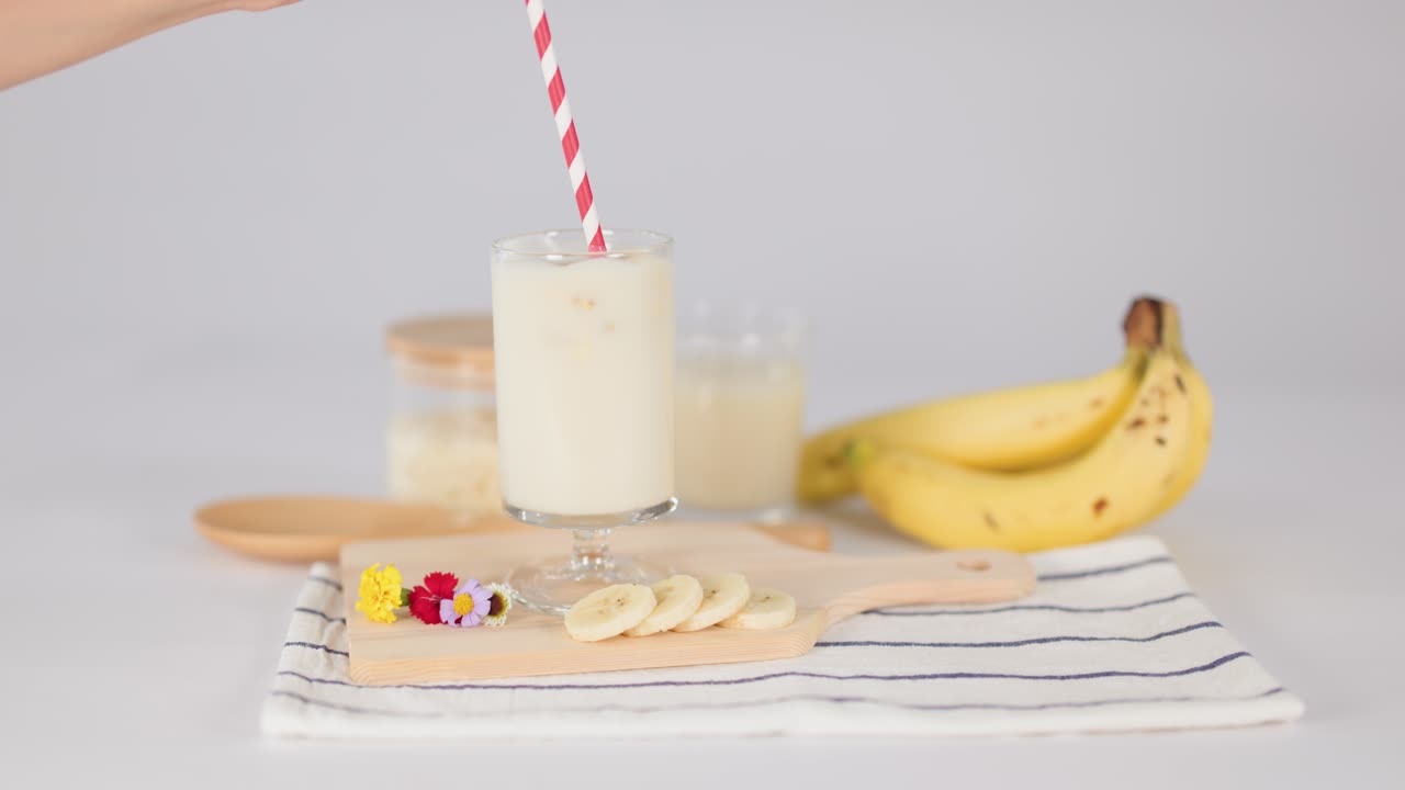 Una mano mescola un cremoso milkshake alla banana con una cannuccia a righe su un tavolo luminoso, banana tagliata in primo piano