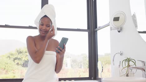 Happy-biracial-woman-with-towel-on-head-washing-her-face-and-using-smartphone,-slow-motion