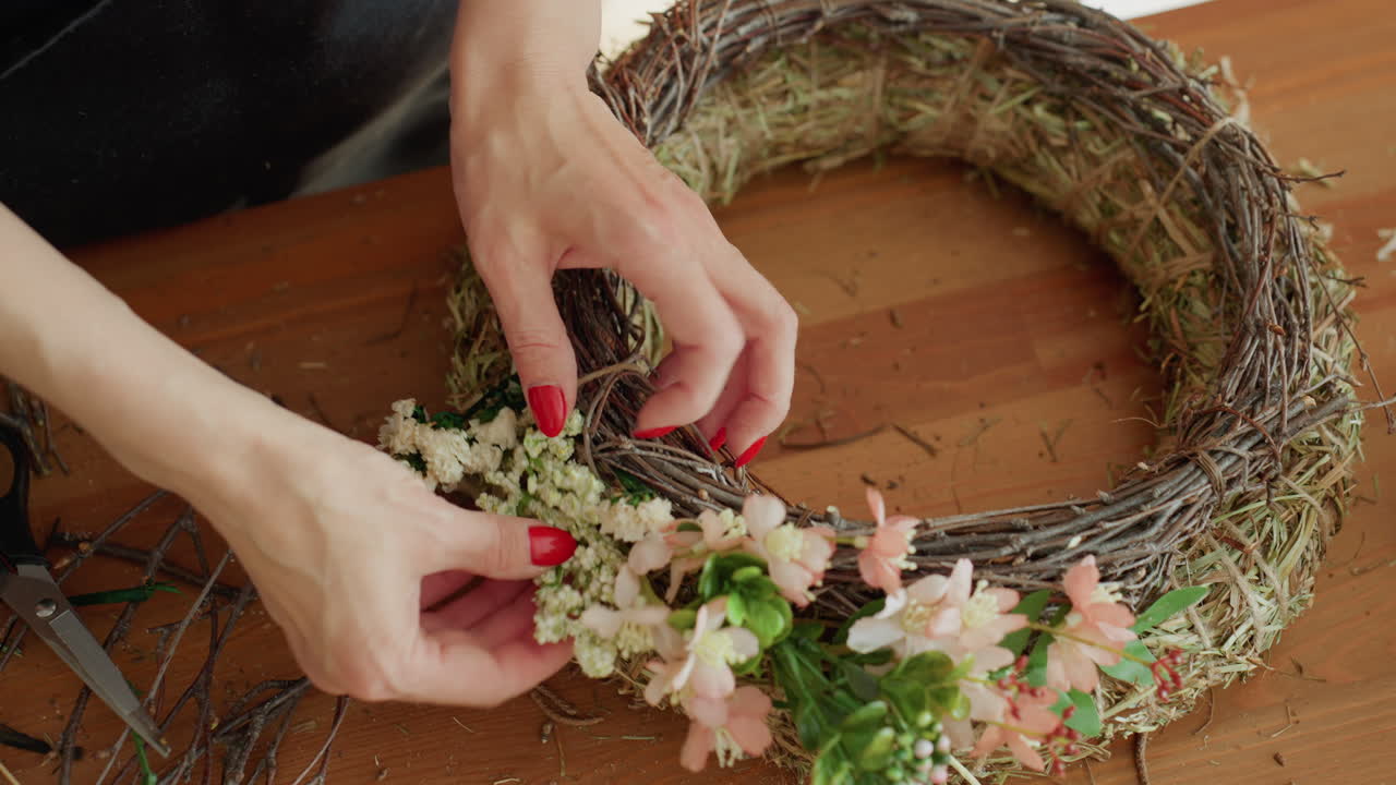 Close up of female hands using scissors to trim small flower stems while decorating rustic wreath with delicate blossoms and greenery, showcasing detailed floral arrangement and handmade crafting process