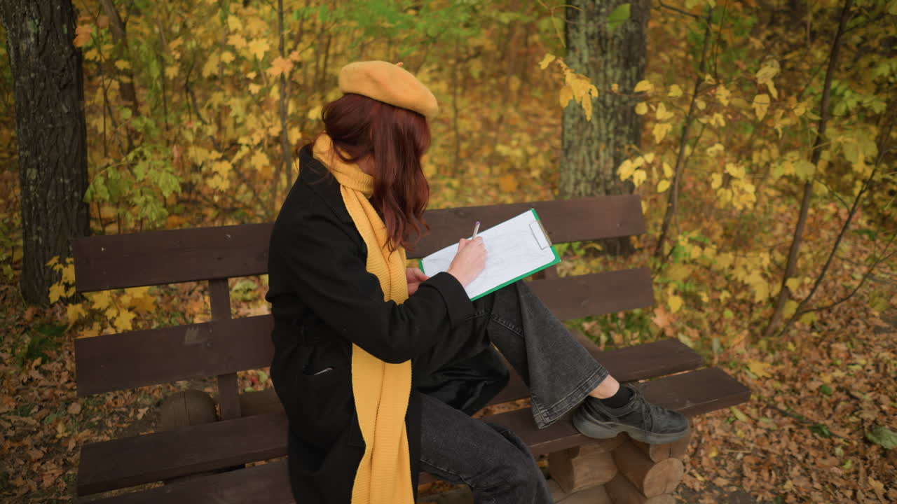 artista profundamente centrada en su dibujo sentada en un banco rodeada de hojas de otoño, con una boina amarilla y abrigo negro en un entorno de bosque sereno