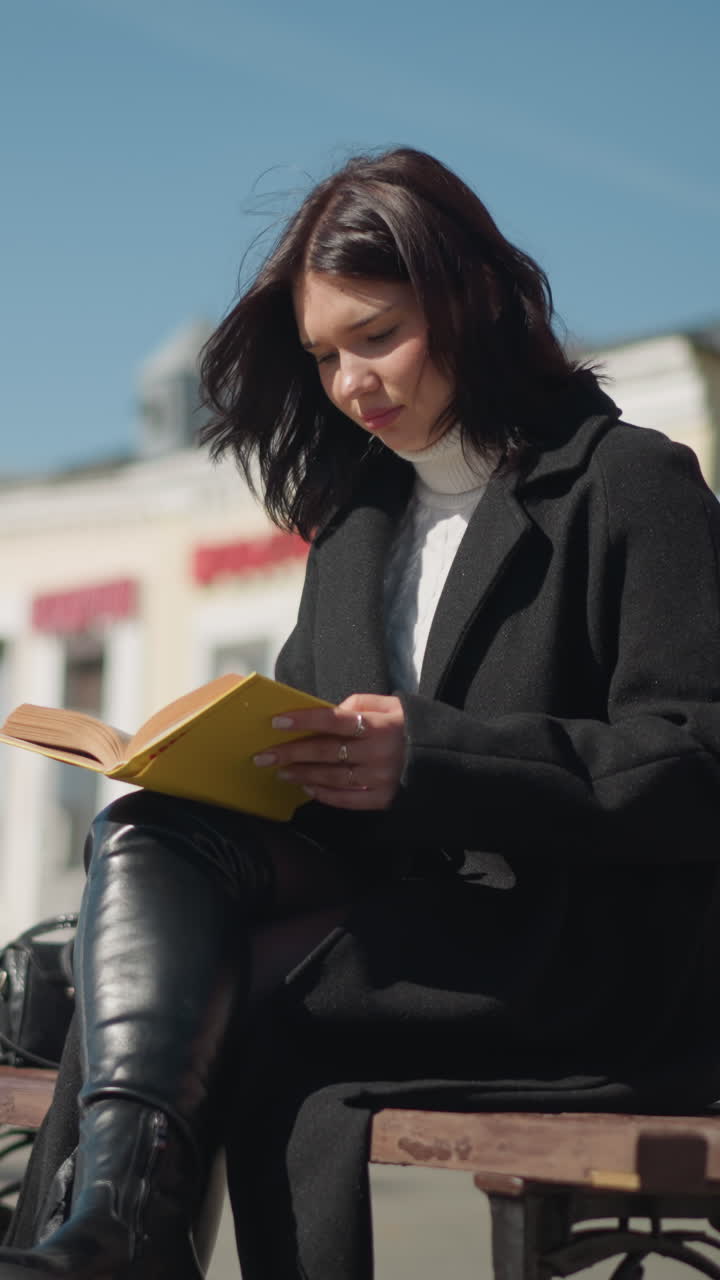 mujer sentada en un banco al aire libre en un día soleado, centrado en la lectura de su libro con páginas de borde amarillo voltea a la siguiente página, su cabello sopla suavemente en la brisa