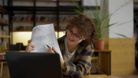 happy girl student with curly hair and glasses demonstrates her work during an online conference in the university library