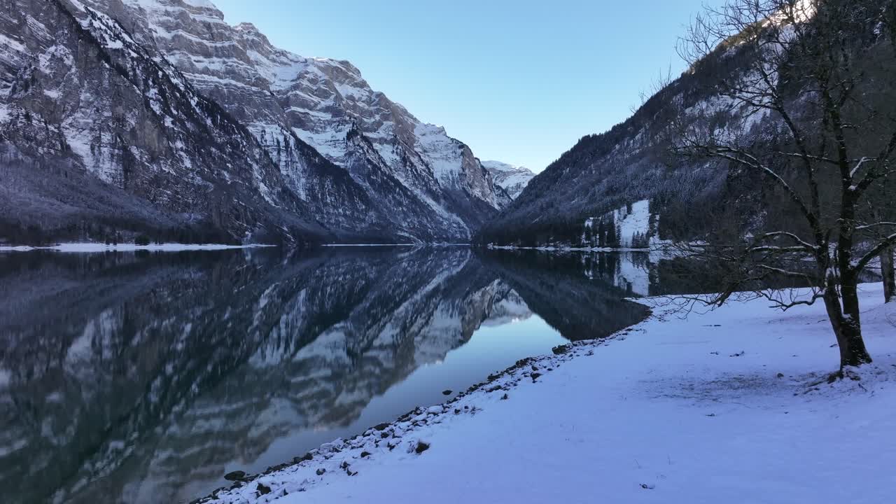 A serene winter scene at Klöntalersee in Glarus, Switzerland. Snow-capped Vorderglärnisch and Vrenelisgärtli peaks frame the tranquil lake, with mirror-like reflections enhancing the landscape.