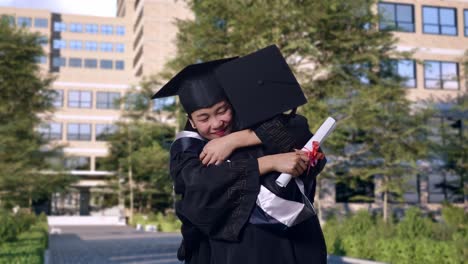 asian woman students graduate in caps and gowns with diplomas smiling and hugging each other in front of a magnificent university building