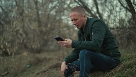 a close view of a man sitting outdoors in a forested area, focused intently on a smartphone in his hand, with a gun in his other hand