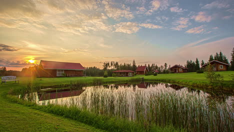 Timelapse-shot-of-a-small-pond-alongside-beautiful-wooden-cottages-with-pine-trees-background-at-sunset