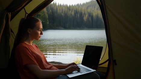 woman working on a laptop inside a tent by a lake in nature