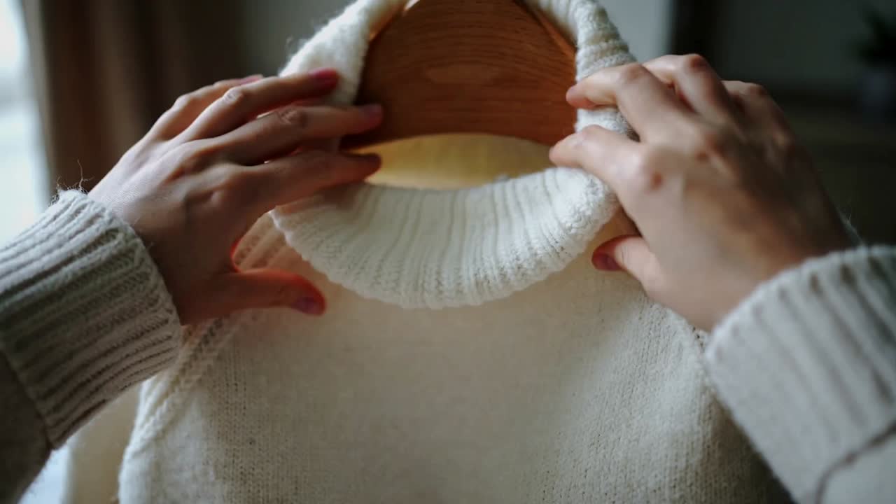 Reaching pink-nailed ringed hands smoothing ivory rollneck on wooden hanger in bedroom showing knit