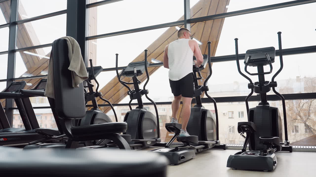 Gym goer exercising on elliptical trainer by large windows in rec center, towel draped over chair, stepping on pedals and adjusting console while checking posture in mirror reflection