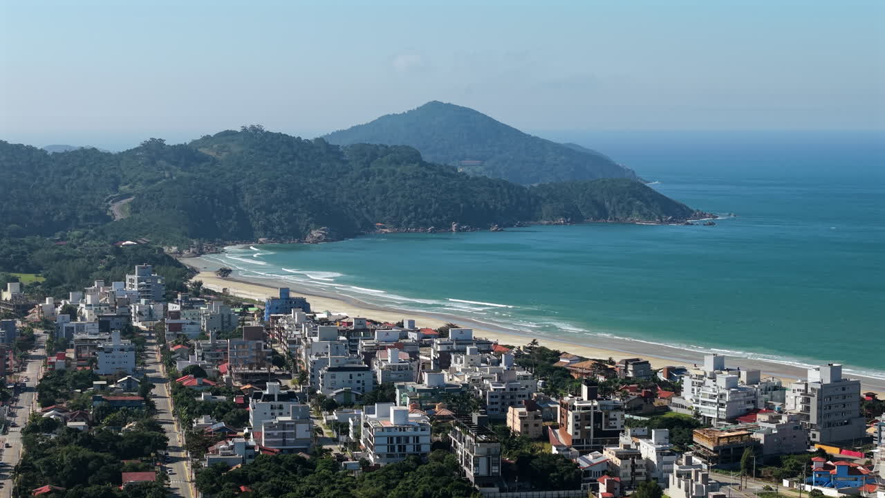 Aerial panorama revealing Mariscal beach, coastal cityscape merging with turquoise Atlantic waters, verdant mountains framing scenic landscape in Santa Catarina, Brazil