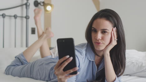 woman wearing pyjamas posing and taking selfies on mobile phone lying on bed