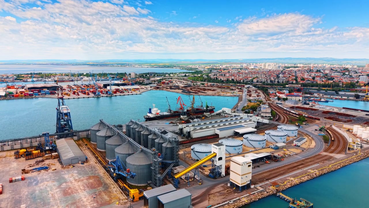 Silos de grano frente al mar. Trabajadores mueven equipos cerca de silos de grano en un puerto concurrido. Los barcos están atracados y los contenedores apilados en tierra.