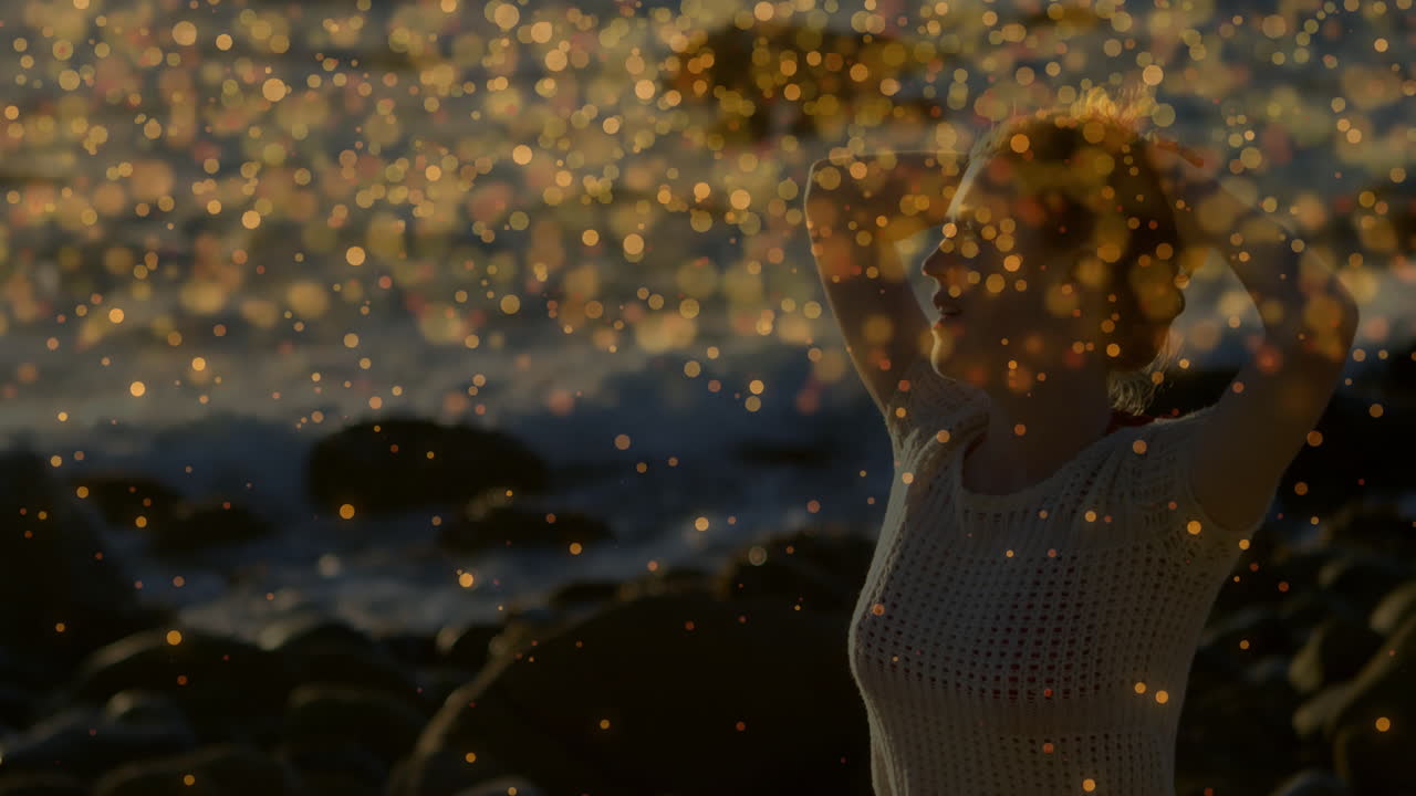 Woman raising arms behind head on rocky beach wearing white crochet top, golden bokeh for health