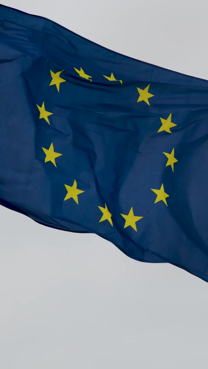 Bandiera europea blu con stelle gialle che sventola vigorosamente in condizioni di cielo coperto e ventoso in riva al mare, vista ravvicinata.