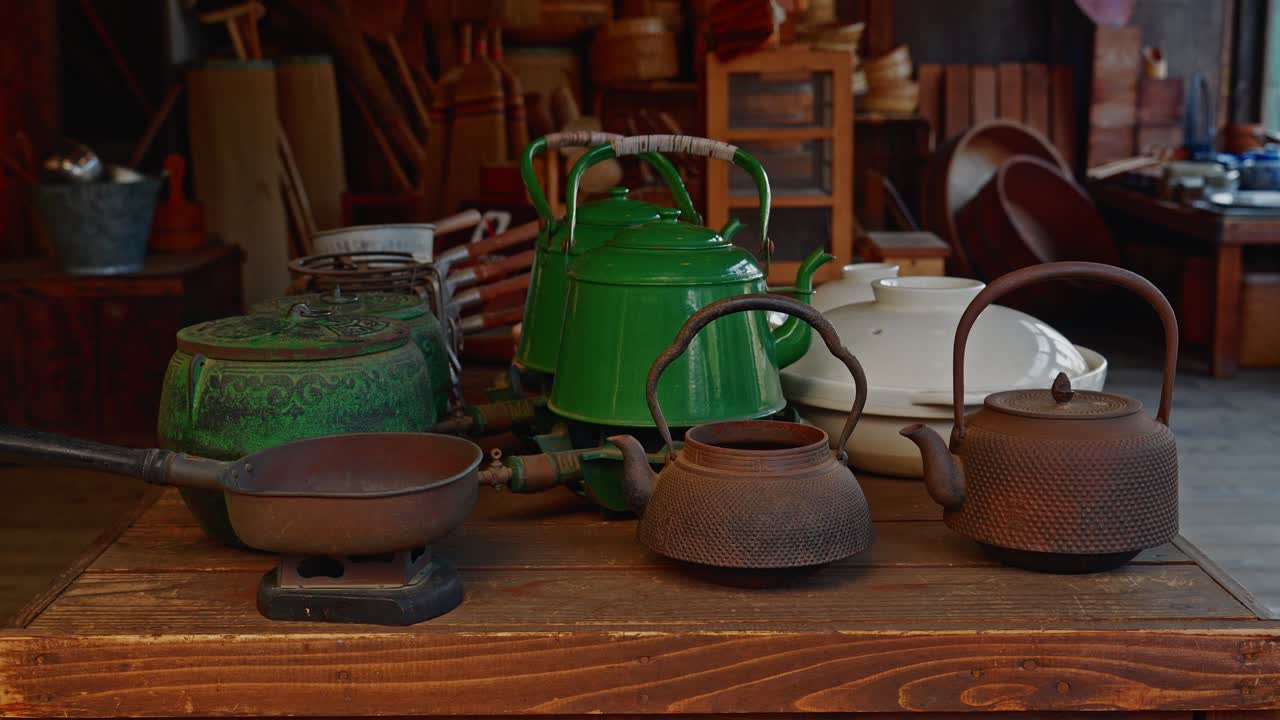 A detailed shot of vintage household items, including rusted iron tetsubin kettles and green enamel pots. Displayed on a wooden table at Edo-Tokyo Open Air Architectural Museum