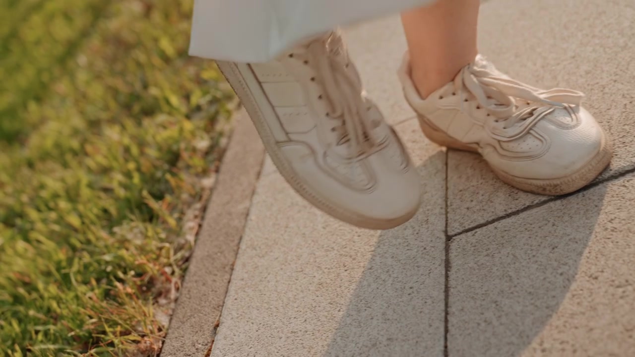 Los pies de una mujer con zapatillas blancas se mueven por un pavimento de piedra soleado, capturando el movimiento rítmico al caminar. El enfoque permanece en sus piernas y zapatos mientras cambian de una baldosa a otra con suavidad.