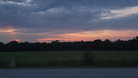 Viajando-Y-Mirando-Por-La-Ventana-Viendo-La-Puesta-De-Sol,-La-Carretera-Y-El-Bosque-Con-Nubes-Oscuras-Y-Cielo-Rojo