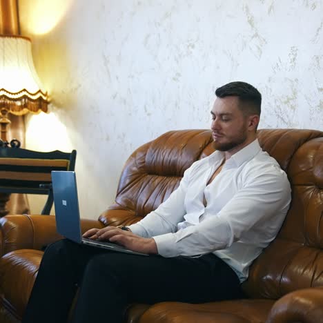 handsome man with a laptop in cosy hotel room. businessman sitting on a couch and working online at business trip. travel suitcase in the room