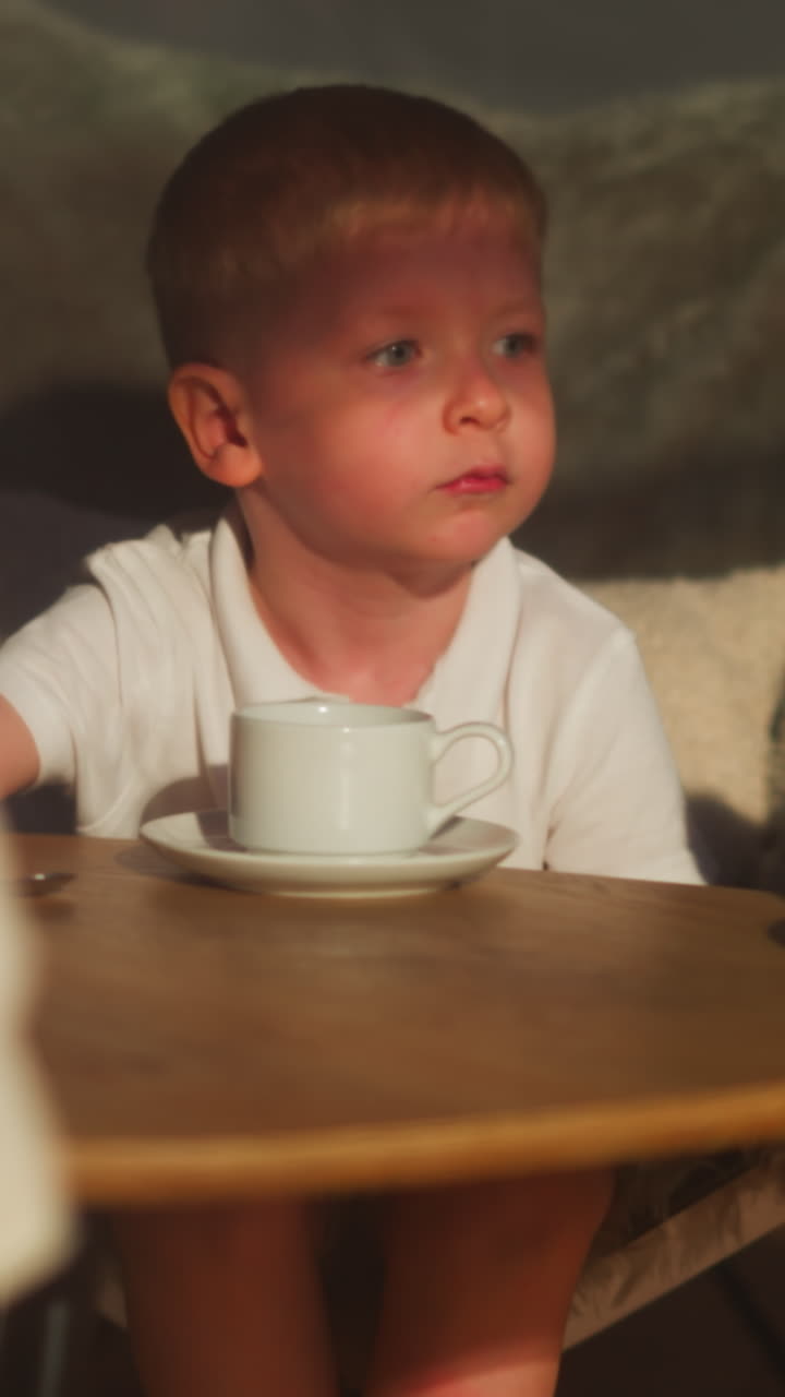 작은 아이는 차 컵과 접시와 함께 커피 테이블에 앉아 있습니다. 조용한 소년은 집에서 여동생과 함께 음료를 마시며 쉬고 있습니다. 사랑스러운 아이는 방에서 포디움에 기대고 있습니다.