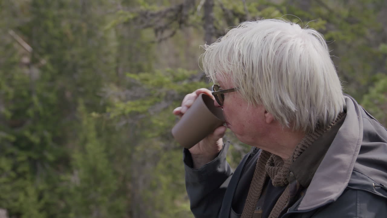 Man with gray hair drinks from cup outdoors in forest while enjoying warm beverage. He is sitting by the view eating.