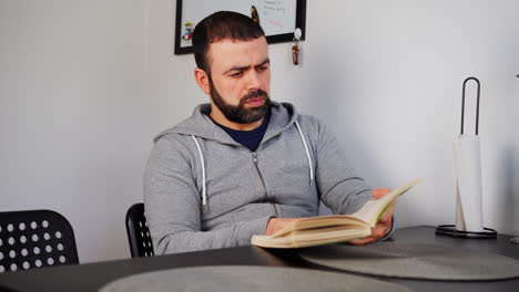 un tipo caucásico del este medio leyendo un libro mientras está sentado en la mesa de comedor