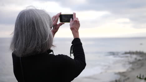 an elderly woman on vacation using her phone to take a picture at the beach over the ocean at sunset slow motion