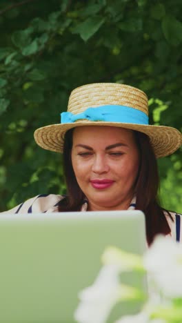 A woman in a straw hat smiles at her laptop while surrounded by greenery, enjoying a peaceful moment outdoors, combining technology with a refreshing nature vibe