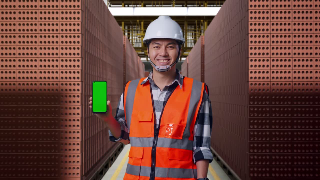 Asian Male Engineer With Safety Helmet Smiling And Showing Green Screen Smartphone To The Camera While Standing With Red Brick Packed in Stacks Are Stored