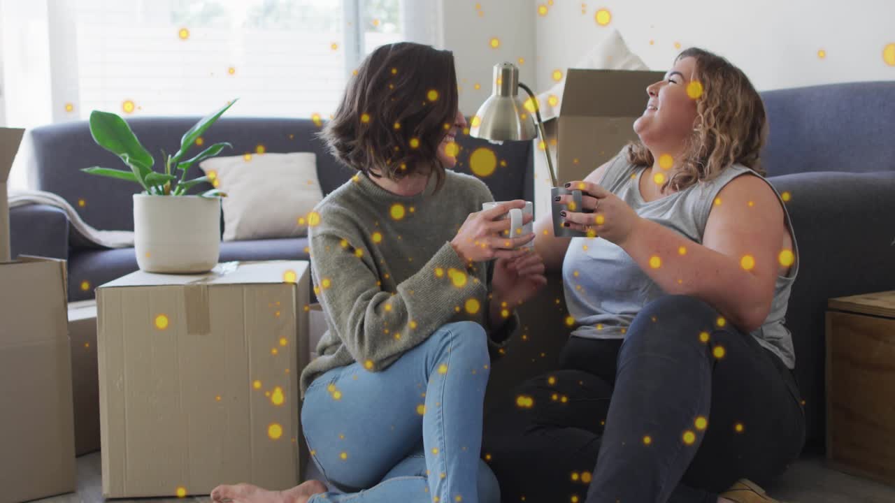 Two women sitting by gray sofa, golden confetti drifting causing mug clinking lifestyle celebration