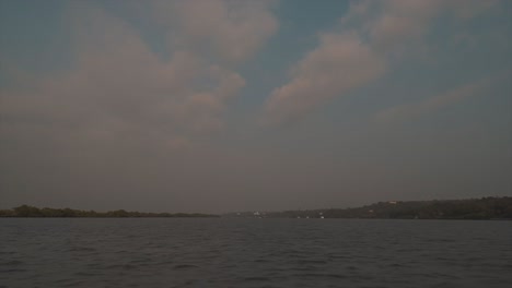 Wide-angle-shot-of-a-lake-on-a-boat-sailing-across-the-lake-with-view-of-calm-water,-trees-and-clouds-in-the-sky