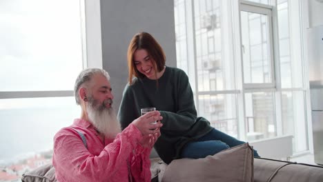 una feliz chica morena en una chaqueta verde oscuro trae un vaso transparente de agua para su padre anciano de cabello gris y una camisa rosa en un apartamento moderno