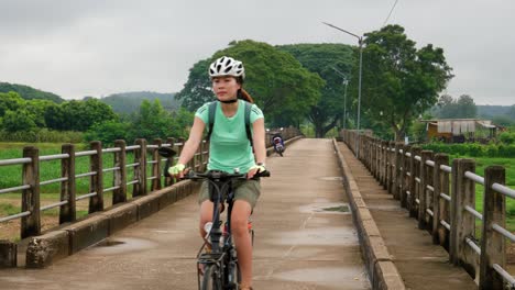 asian adult woman riding and pedaling bike towards camera on bridge in thailand