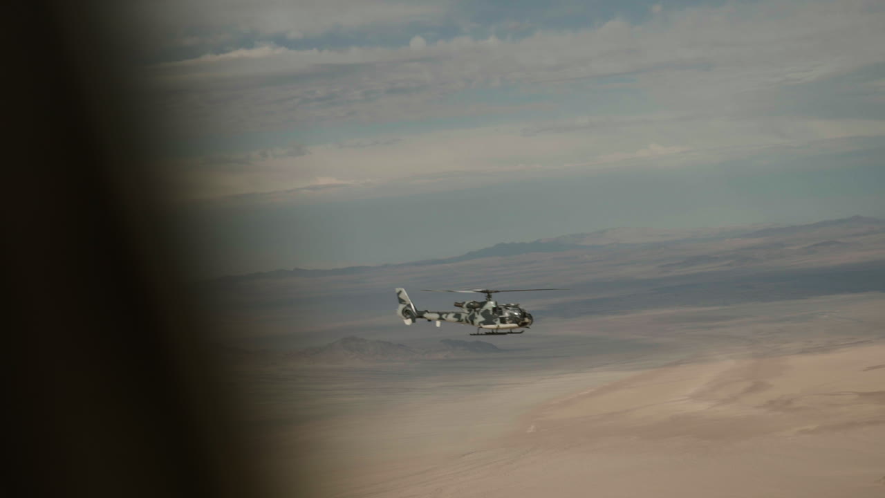 A gazelle helicopter is seen gracefully flying across a vast desert landscape. The sky is clear, showcasing the natural beauty of the terrain beneath the flying aircraft.
