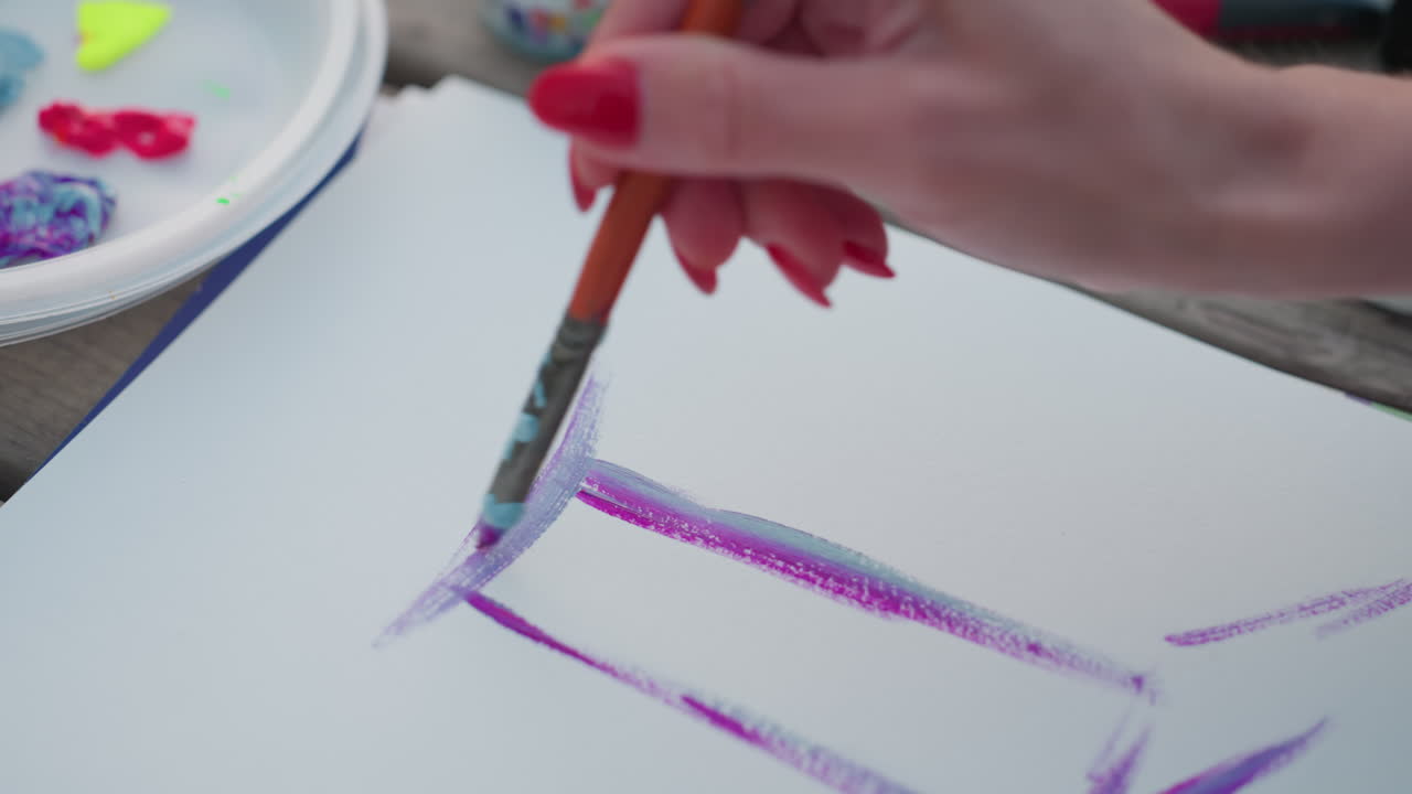 close up shot of lady hand blending vibrant violet and blue watercolor strokes on white paper with soft golden light highlighting paint palette and pastel jar on wooden bench by riverbank at sunset