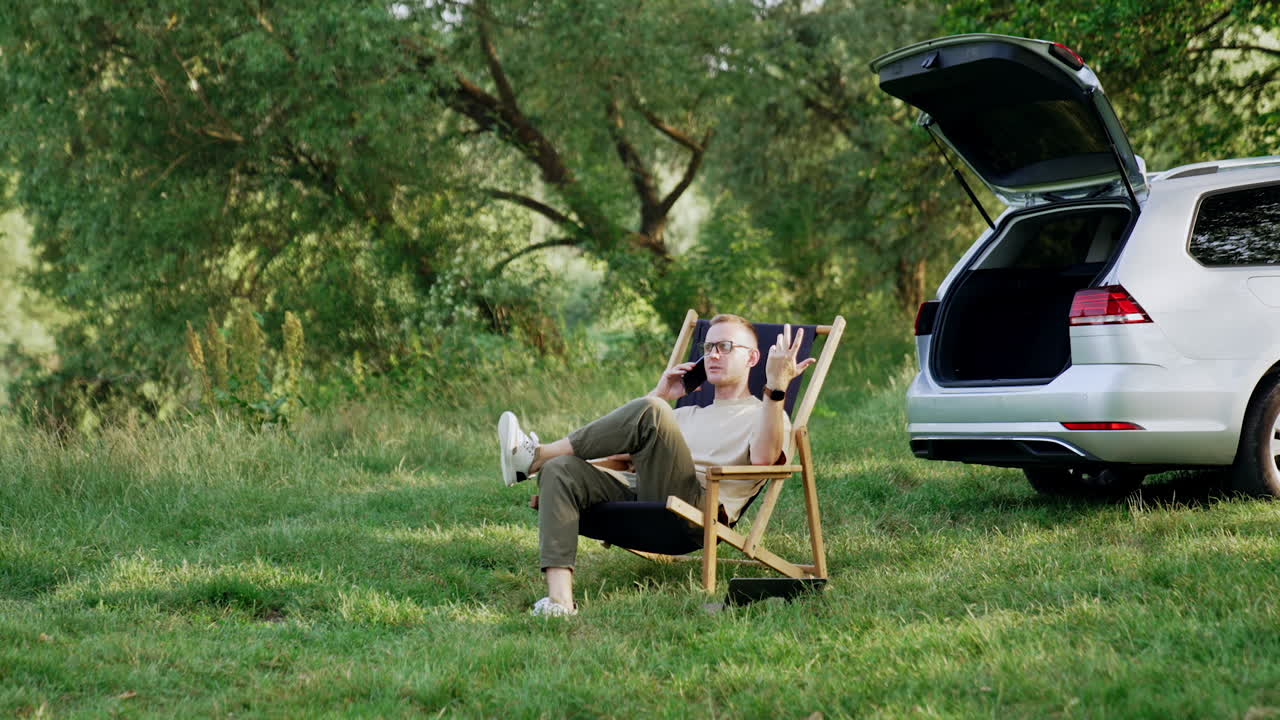 Relaxed male speaks on the phone sitting in the folding chair. Remote work conversation in the woods.