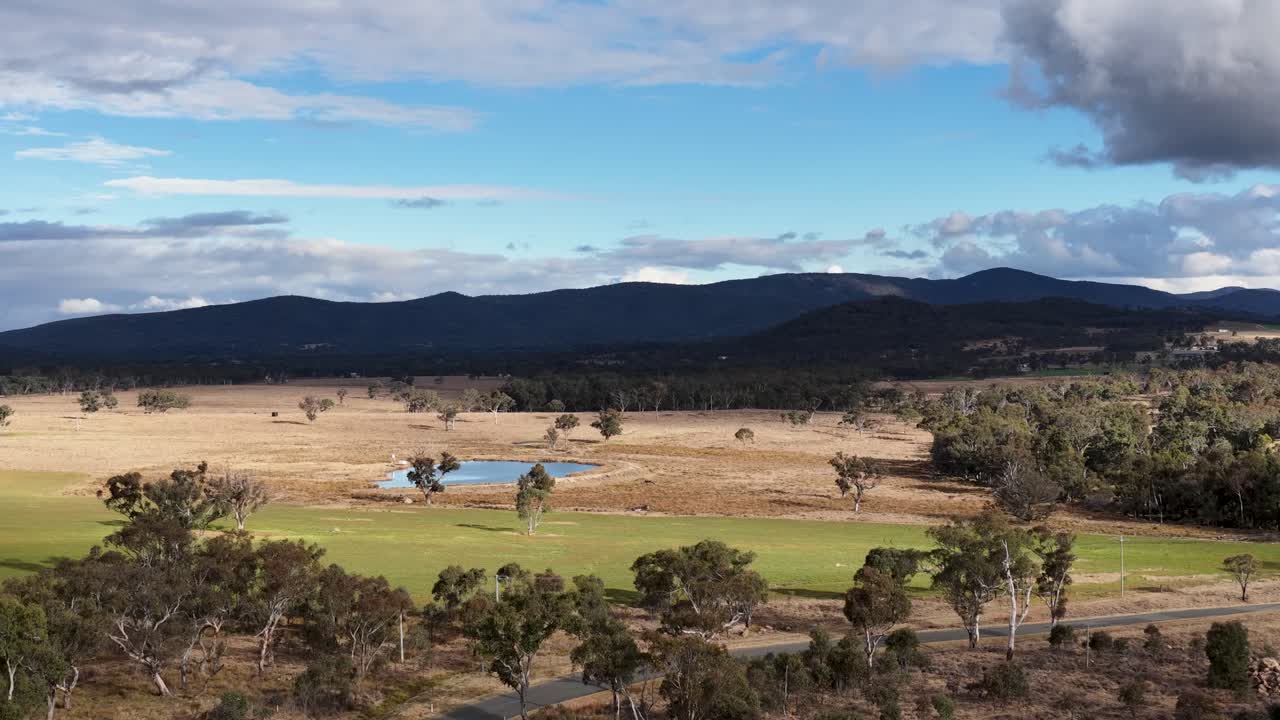 Aerial footage pans smoothly across rural Stanthorpe, Queensland, revealing fields, scattered trees, distant mountains, and dramatic clouds under soft daylight